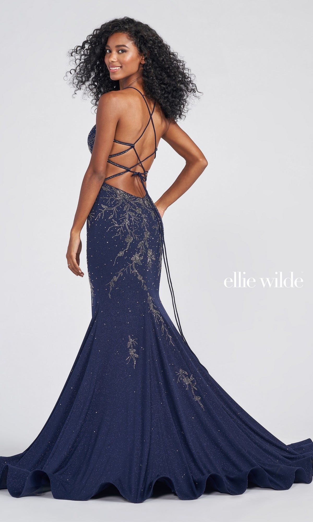 A woman poses with her back to the camera in the Ellie Wilde EW122028 Sparkle Jersey Formal Dress, a navy blue mermaid gown with gold embellishments, crisscross straps, and a backless design.